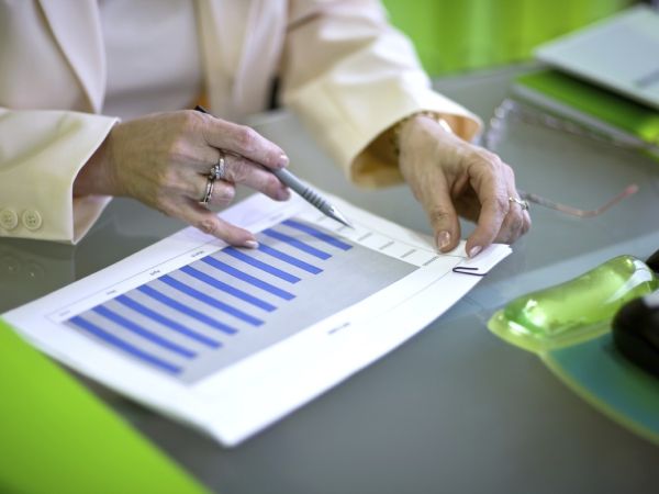 Eine Geschäftsfrau sitz an einem Schreibtisch mit einem Kulli in der rechten Hand und überprüft Zahlen in einem Diagramm, daß auf einem Deckblatt eines mit einer Büroklammer zusammen-gehefteten Blätterstabels abgebildet ist. Sie hebt diesen Blätterstabel